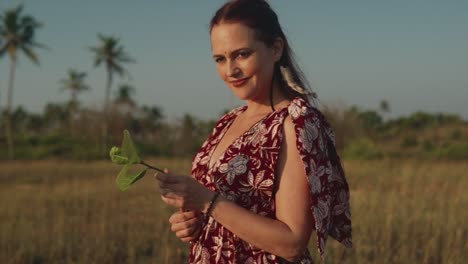 Hermosa-Mujer-Con-Un-Vestido-Estampado-Floral-Parada-En-Un-Campo-Sosteniendo-Una-Hoja-En-Sus-Manos-Y-Sonriendo-A-La-Cámara