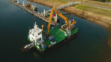 Tiro-De-Dron-De-Retroceso-Desde-Un-Barco-De-Dragado-Con-Excavadora-De-Grúa-Amarilla-En-Un-Pequeño-Puerto