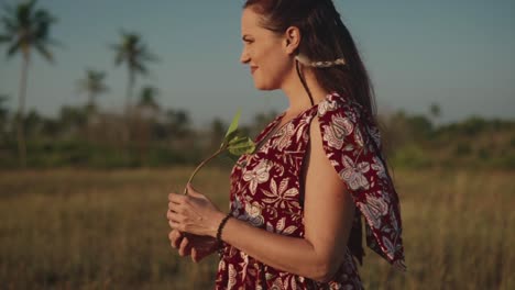 Dame-In-Einem-Kleid,-Die-Auf-Einem-Feld-Steht-Und-Lächelt,-Während-Sie-Eine-Grüne-Blume-In-Ihren-Händen-Hält