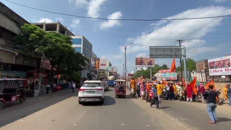 Toma-Pov-De-Una-Carretera-De-Dos-Vías-Junto-A-Un-Puesto-De-Autos-Y-Una-Procesión-Que-Pasa-Por-Dhanbad,-India,-En-Un-Día-Soleado