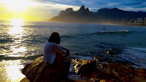 Mujer-Sentada-En-Las-Rocas-En-La-Playa-De-Arpoador-Al-Atardecer-Con-Las-Icónicas-Montañas-De-Río-De-Janeiro-En-Silueta