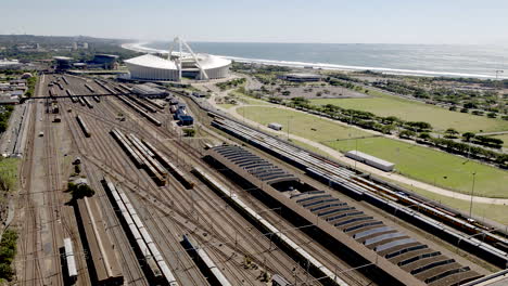 Toma-Aérea-Del-Tren-De-Pasajeros-Que-Sale-De-La-Estación-De-Tren-De-Durban-En-Sudáfrica-Con-El-Estadio-De-Fútbol-Moses-Mabhida-Y-El-Océano-índico-En-Segundo-Plano