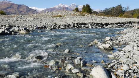 Low-angle-close-up-of-flowing-river-between-stones-and-rocks-during-Glacier-Burn-Track-in-new-Zealand---Snowy-Mountains-in-background