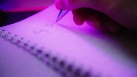 Close-up-shot-of-a-hand-writing-dreamy-motivated-words-on-a-note-paper-with-his-pen-in-dark-purple-gaming-room