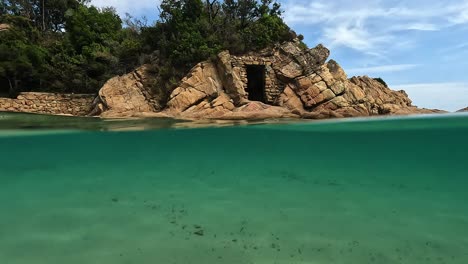 Half-underwater-view-of-old-seafront-passage-door-carved-into-rock-of-Canella-idyllic-beach-in-Corsica-island,-France