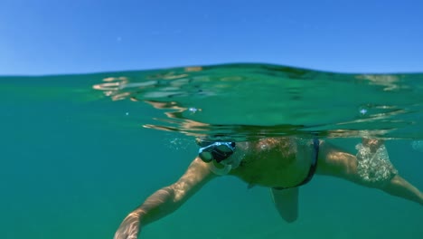 Half-underwater-view-of-adult-man-snorkeler-diving-to-see-and-touch-Rhizostoma-Pulmo-jellyfish-medusa-while-holding-bag-for-clam-and-mussel-fishing
