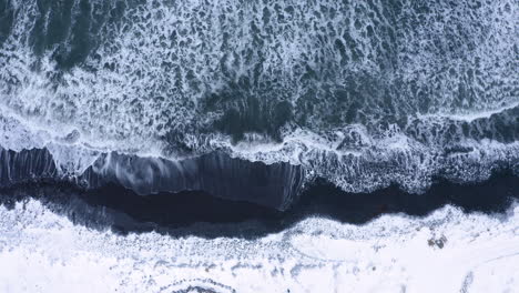 Drone-top-down-shot-of-Arctic-ocean-waves-in-winter-splashing-and-reaching-black-volcanic-beach-in-Iceland