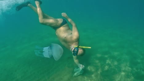 Half-underwater-view-of-adult-man-snorkeler-diving-to-see-Rhizostoma-Pulmo-jellyfish-medusa-while-holding-bag-for-clam-and-mussel-fishing