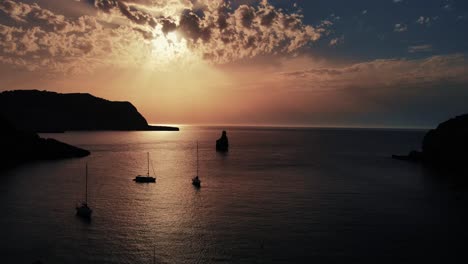 coastal-sunset-blasting-through-clouds-aerial-view-in-Cala-Benirras,-Ibiza-overlooking-yachts-and-boats-on-the-coast-sliding-forward