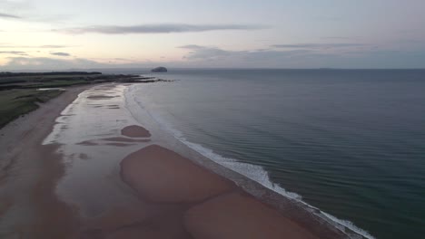 Drone-footage-slowly-descending-towards-a-long-sandy-beach-as-a-sunset-reflects-in-tidal-pools-and-the-tide-and-waves-gently-lap-the-shore-of-the-beach