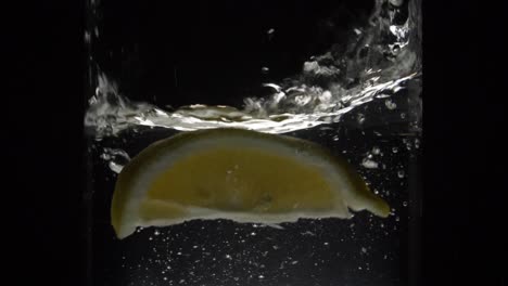 Slow-Motion-Of-A-Lemon-Slice-Falling-Into-A-Glass-Of-Water-Isolated-In-Black-Background---medium-shot,-180fps