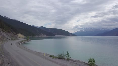 A-motorcyclist-traveler-driving-along-curved-road-at-the-side-of-a-stunning-canadian-lake-with-mountain-range-on-sides-on-a-cloudy-day,-locked-down-panoramic-shot