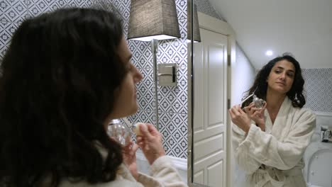 Young-beautiful-middle-eastern-woman-wearing-a-bathrobe-and-standing-in-a-fancy-bathroom,-spraying-luxury-perfume-out-of-a-vintage-glass-bottle-with-pump
