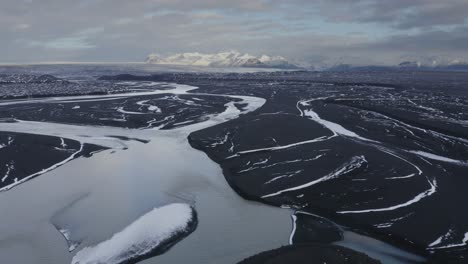 Aerial-flyover-glacial-Su-la-River-Delta-and-snowy-mountains-in-background-during-cloudy-day-on-Iceland