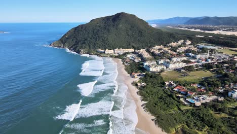 Aerial-drone-scene-of-paradisiacal-beach-view-of-the-high-Brazilian-coastline-with-mountains-sand-ocean-horizon-buildings-beach-houses-nature-and-resort-Santinho-beach-Florianópolis