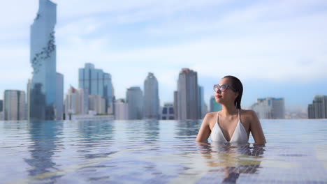 Attractive-asian-female-in-infinity-rooftop-pool-of-luxury-Bangkok-hotel-enjoying-sun,-cityscape-in-background