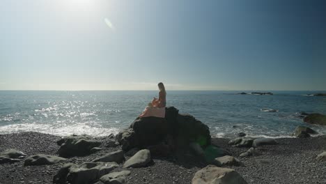 Woman-having-moment-alone-writing-in-her-journal-while-sitting-on-rock-at-sea,-summer-day
