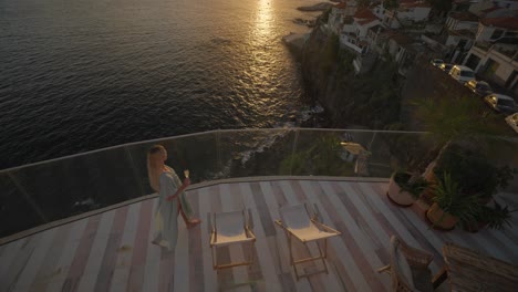 Woman-in-elegant-dress-with-glass-of-champagne-on-luxury-balcony-with-stunning-sunset-view-of-Funchal,-Madeira