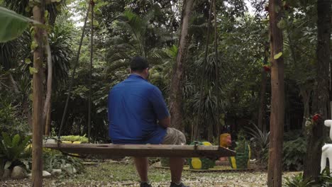 A-Shot-of-a-well-built-Caucasian-male-sitting-on-a-swing,-swaying-back-and-forth-as-he-sits-alone-thinking-in-the-middle-of-a-tropical-forest-garden-in-Kanchanaburi-Thailand