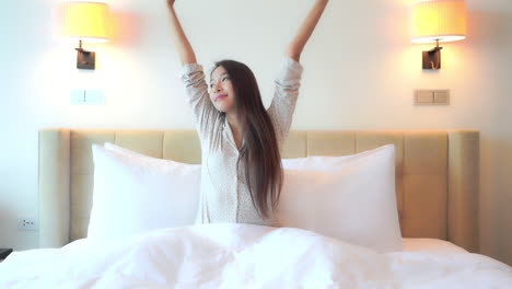 Pretty-Asian-woman-smiles-and-stretches-arms-white-sitting-in-bed-with-morning-light-shining-through-window-out-of-frame
