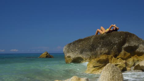 Woman-in-bikini-sunbathe-lying-on-granite-cliff-washed-by-crystal-turquoise-sea-water-on-tranquil-beach