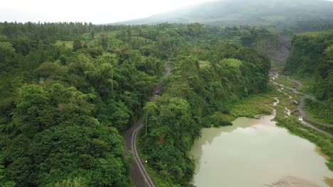 Aerial-tracking-shot-of-trucks-driving-on-sand-mine-road-between-forest-trees-of-Merapi-Volcano