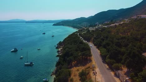 Aerial-shot-of-coastal-road,-moving-cars,-blue-ocean-and-small-city-in-Corfu,-Greece