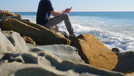 Ein-Mann,-Der-Sich-Hinsetzt-Und-Auf-Seinem-Smartphone-Eine-SMS-Schreibt,-Während-Die-Wellen-Des-Kalifornischen-Ozeans-In-Zeitlupe-Gegen-Die-Felsige-Küste-Prallen-Und-Sprühen