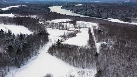 Luftaufnahmen-Fliegen-Hoch-über-Mehrere-Schneebedeckte-Felder-Zu-Einem-Teilweise-Zugefrorenen-Teich-In-Einem-Winterwald