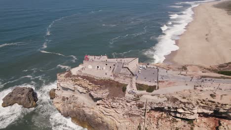 Nazare-lighthouse-and-Saint-Michael-the-Archangel-fort-perched-atop-of-rocky-cliff,-aerial-pan-shot