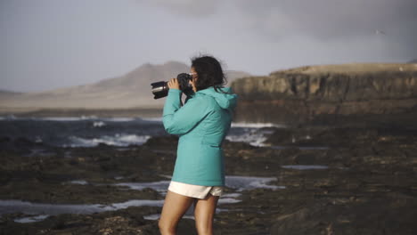 Fotógrafo-Creativo-Independiente-Parado-En-El-Acantilado-Del-Océano-Durante-Un-Día-Ventoso-Con-Una-Chaqueta-De-Moda-Tomando-Fotos-Del-Paisaje-Marino-Natural-En-Fuerteventura,-España,-Islas-Canarias