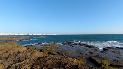 Waves-breaking-at-stone-reef-with-ocean-and-city-in-distance