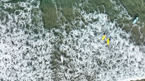 Top-down-aerial-shot-of-surfers-wading-through-sea-foam-and-entering-the-ocean-to-surf-waves
