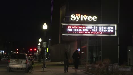 Two-people-walking-past-a-music-venue-marquee-at-night