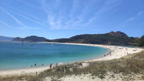 Gente-Disfrutando-De-La-Tarde-En-La-Playa-Con-Dunas-De-Arena,-Barcos-Anclados-Y-Rastros-De-Aviones-En-El-Cielo-Soleado,-Tiro-Panorámico-Bloueado,-Islas-Cíes,-Pontevedra,-Galicia,-España