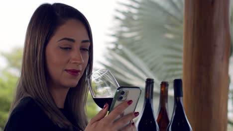 Toma-Cinematográfica-De-Una-Dama-Sentada-En-Un-Jardín-Tropical-Con-Una-Copa-De-Vino-Tinto-En-La-Mano-Mirando-Su-Teléfono-Inteligente---Cierre-A-Cámara-Lenta