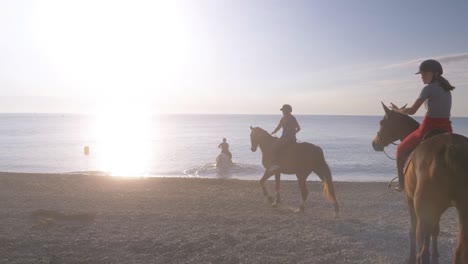 Gruppe-Von-Touristen,-Die-Am-Sandstrand-Reiten-Und-Bei-Sonnenuntergang-Ins-Wasser-Tauchen---Langsamer-Vorwärtsschuss