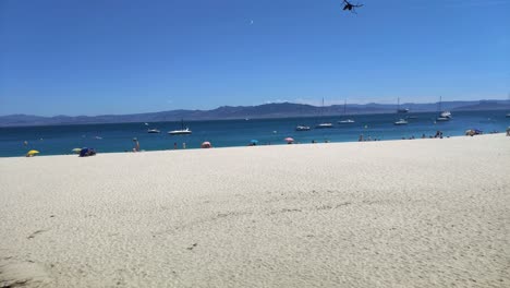 people-enjoying-afternoon-on-the-beach-with-sand-dune,-jetty-with-restaurant,-anchored-boats,-day-with-sunny-sky,-panoramic-shot-turning-right,-Cíes-Islands,-Pontevedra,-Galicia,-Spain