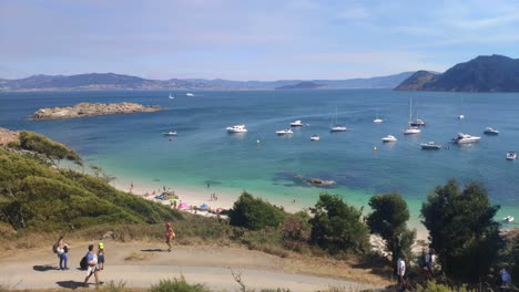 people-walking-in-nature-and-the-sea-in-the-background-with-anchored-sailboats,-the-island-in-the-background,-sunny-day,-panoramic-overhead-shot-turning-right,-Cíes-Islands,-Pontevedra,-Galicia,-Spain