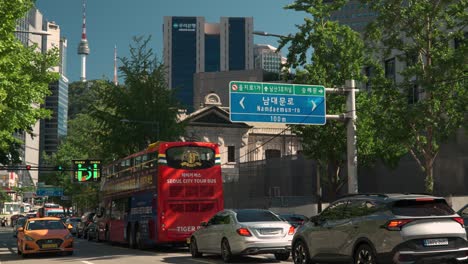 Double-deck-red-Seoul-city-tour-bus-stuck-in-a-traffic-jam-at-narrow-old-town-street-on-Sogong-ro-road-with-a-view-of-iconic-N-Seoul-Tower-or-Namsan-Tower-in-background