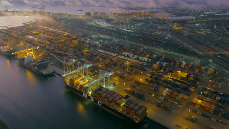 The-Port-of-Oakland-at-nighttime-with-cargo-ships-and-cranes-at-the-dock-and-glowing,-dreamy-lights-in-the-foggy-dusk---aerial-view