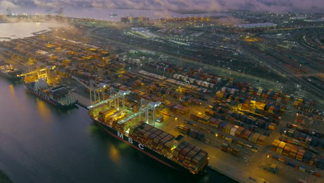 El-Puerto-De-Oakland-Por-La-Noche-Con-Buques-De-Carga-Y-Grúas-En-El-Muelle-Y-Luces-Brillantes-Y-Soñadoras-En-El-Oscuro-Atardecer---Vista-Aérea