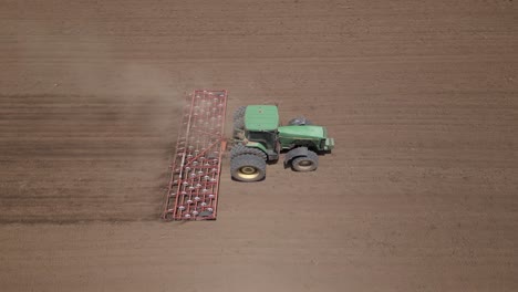 Aerial-dolly-shot-of-green-tractor-pulling-tine-harrow-in-field-soil