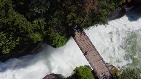 People-crossing-spectacular-bridge-over-Strandaelvi-river-in-Voss-Norway---Summer-birdseye-view-with-lush-forest-on-sides-and-powerful-river-beneath-bridge