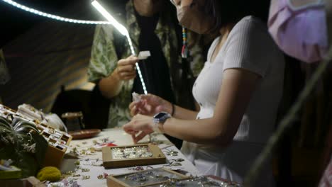 Mujer-Asiática-Con-Máscara-Facial-Mientras-Busca-Joyas-En-El-Mercado-Nocturno-Al-Aire-Libre,-Filmada-A-Mano-Con-Una-Inclinación-Lenta-Hacia-Abajo