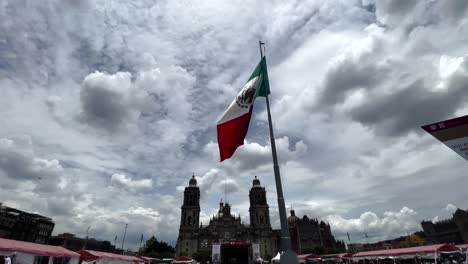 Toma-De-La-Bandera-De-México-Ondeando-Con-La-Vista-Completa-Del-Zócalo-En-El-Fondo-Al-Atardecer-Frente-A-La-Catedral-Metropolitana