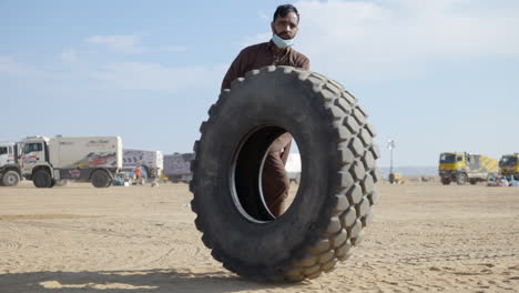 Hombre-árabe-Saudita-Empujando-Un-Enorme-Neumático-A-Través-Del-Desierto-Arenoso-En-El-Rally-Dakar