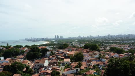 Dolly-in-aerial-drone-shot-of-the-stunning-coastline-of-the-historic-city-of-Olinda-with-small-local-homes-below-facing-towards-the-large-modern-city-of-Recife-in-Pernambuco,-Brazil