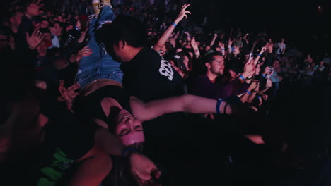 Los-Espectadores-Se-Amontonan-Frente-Al-Escenario-Animando-A-Una-Joven-En-Brazos-En-Un-Concierto-De-Música-Rock,-Los-Agentes-De-Seguridad-Supervisan,-Dentro-De-La-Arena-Con-Coloridos-Focos-Intermitentes