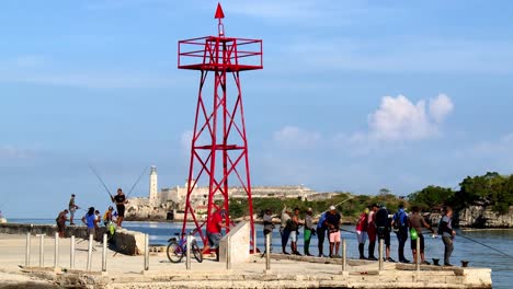 People-fishing-from-the-dock-with-the-Faro-Castillo-del-Morro-lighthouse-in-the-background-in-Havana,-Cuba